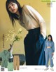 A woman in a beige shirt and blue pants standing next to a vase of flowers.